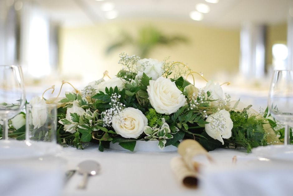 A floral arrangement featuring a centerpiece of white roses, baby's breath, and variegated greenery placed on a dining table. The bouquet is styled in a low, lush arrangement with fresh, vibrant petal
