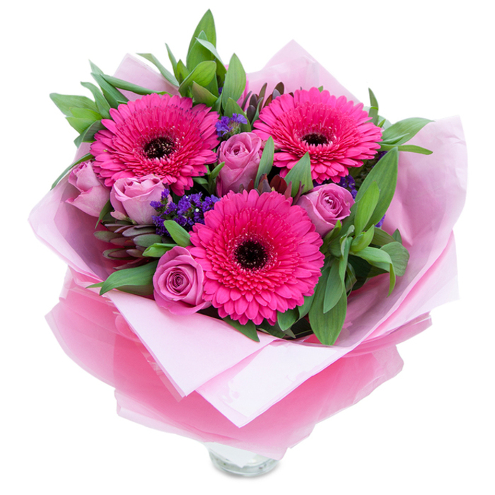 An intimate, graceful bouquet that balances playfulness and refinement, described as if placed near a sunlit window overlooking Calverley Park. At the heart sit three fuchsia gerberas, each with perfectly radiating petals and dark, prominent centres that create a striking contrast against the softer tones. Softer lavender-pink roses nestle close to those gerberas, their petals still snugly rolled in a fresh, budding form that promises to open with time and light, releasing a subtle, sweet rose scent. Dotted throughout are tiny clusters of deep purple statice, lending a suggestion of wildflower spontaneity and delicate texture among the larger blooms. The greenery is generous and tactile - broad, glossy leaves like salal alongside the slender grey-green hints of eucalyptus - which enrich the bouquet with cool, natural tones and a slightly dewy visual freshness. The arrangement is wrapped for giving: an inner sheet of matte pale pink tissue supports the flowers, while an outer layer of translucent fuchsia-pink cellophane adds a soft sheen and holds the theme together. The glass vase rim peeks out at the base, catching a sliver of daylight and signalling that the bouquet is ready for immediate display on a mantlepiece or kitchen island. This composition conveys warmth, celebration, and gentle affection - a perfect choice for anniversaries, a thoughtful birthday surprise, or a simple gesture to brighten someone's day in Royal Tunbridge Wells. The combination of bold gerberas and tender roses reflects careful floral craftsmanship and attention to seasonal freshness.
