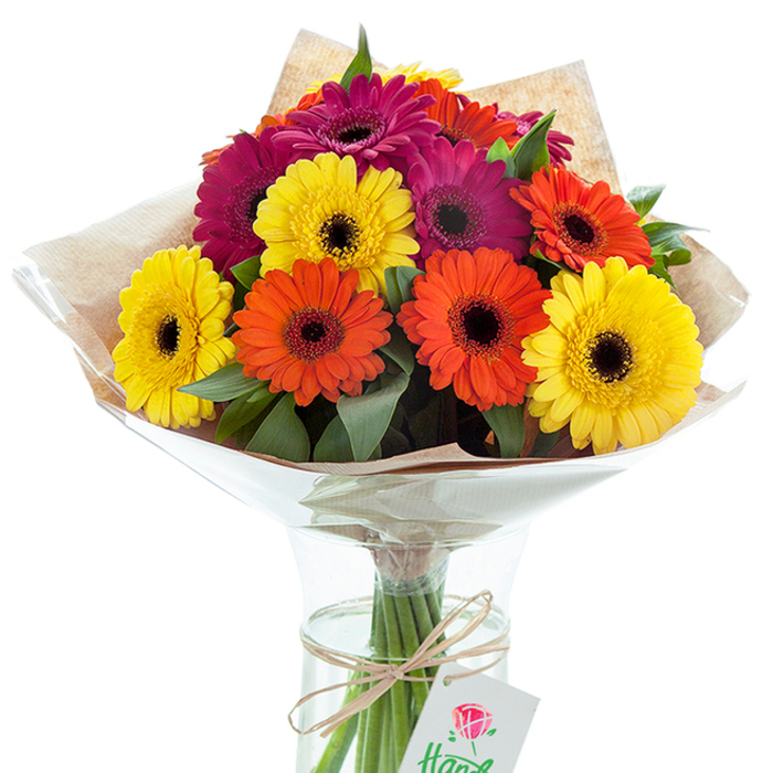 An artfully crafted bouquet of Gerbera daisies, bursting with life and colour, stands as the centerpiece in a sparkling glass vase filled with clear water. The bold hues-cheerful yellow, vibrant orange, and sultry magenta-each encircle a dramatic dark core, drawing attention and evoking pure summer exuberance. Lush green leaves interweave between blooms for texture and fullness, echoing the lush landscapes found around Dunorlan Park in Tunbridge Wells. A double wrapping of subtly textured kraft paper and gleaming cellophane flares lightly at the top, setting off the cluster's elegant, natural form. At the base, a piece of rustic twine is tied with precision, suspending a simple white gift tag decorated with a red rose illustration-a subtle suggestion of heartfelt gifting or special celebration. The clear vase's shape showcases the healthy stems, their green lines upright and perfectly trimmed, promising long-lasting beauty for any home or special event. This floral arrangement, illuminated by studio lighting, captures the essence of joy and giving. Whether delivered by Flowers Tunbridge Wells for a birthday, anniversary, or spontaneous display, the bouquet's expert composition and harmonious colour story speak of care, artistry, and the vibrant bloom of life in Tunbridge Wells.